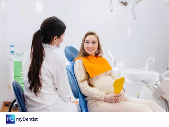 Mujer embarazada con babero naranja y espejo amarillo sentada en la sala del dentista hablando con ella y el logo myDentist